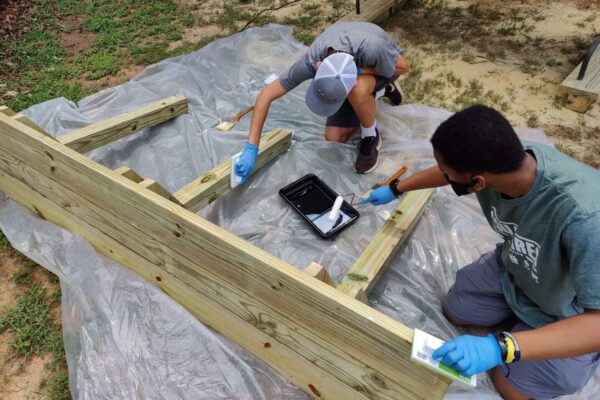 Two people painting a wooden structure with blue gloves.