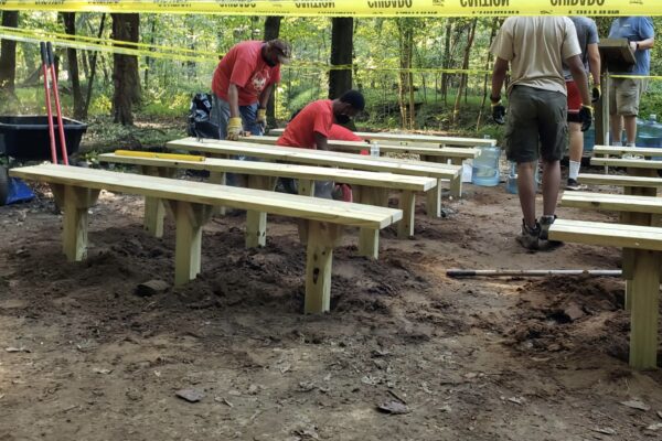 A group of people standing around benches in the woods.