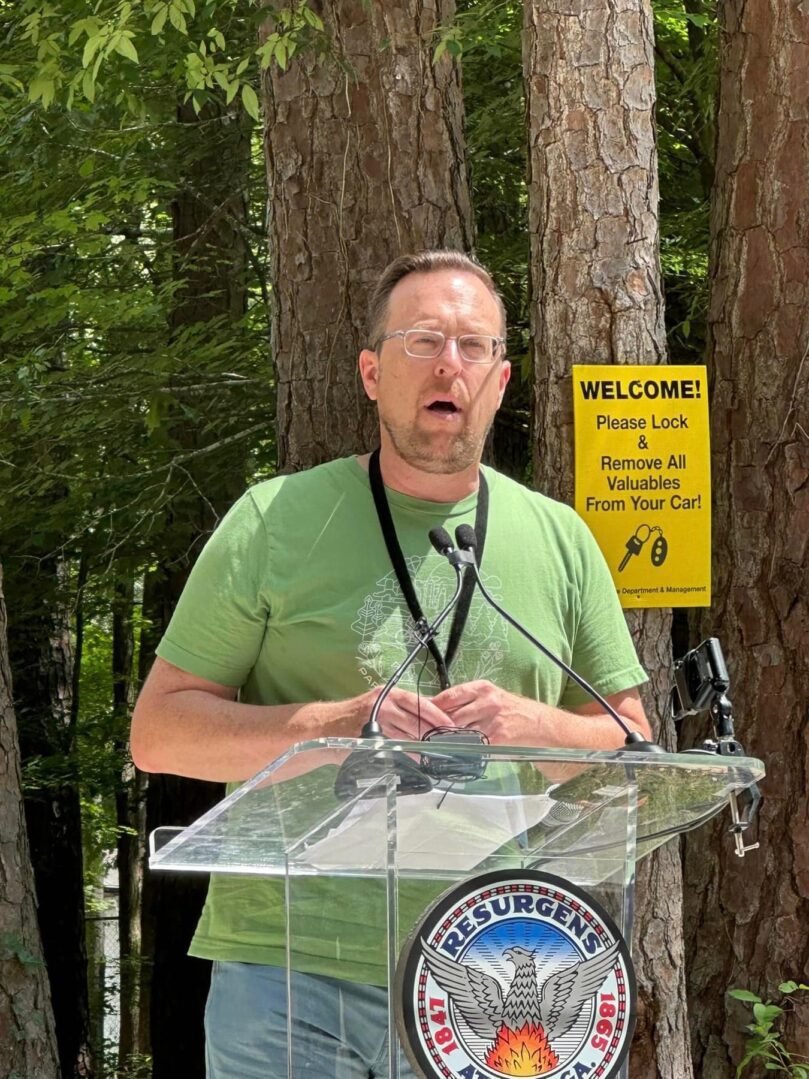 A man in green shirt standing at podium near trees.