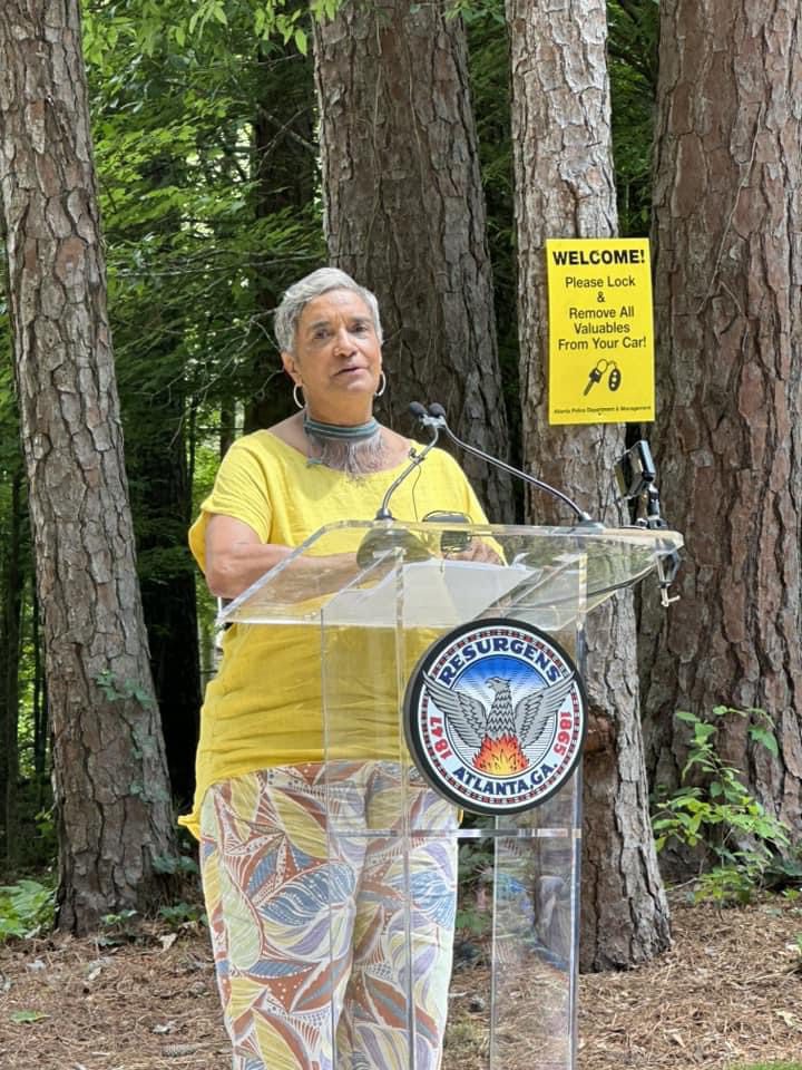 A woman standing at a podium in front of trees.