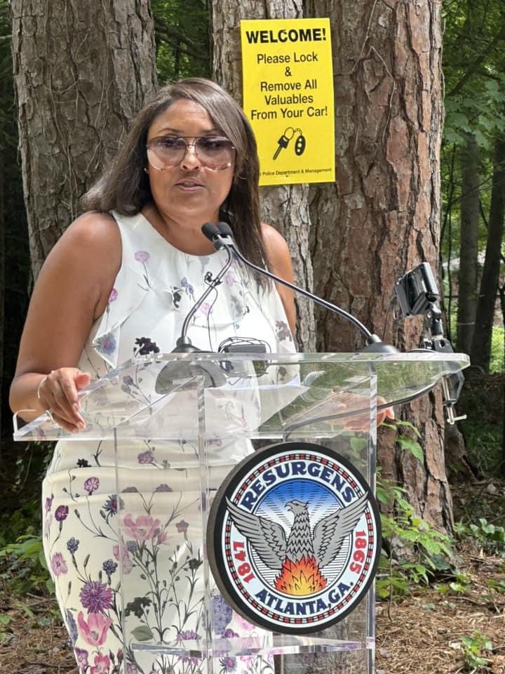 A woman standing at a podium in front of trees.